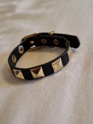 Svart skinnarmband med nitar - Säljer ett svart armband i skinn med fyrkantiga guldfärgade nitar runt om. Armbandet har justerbar spänne och ger en cool och edgy vibe till din outfit. Perfekt för dig som gillar detaljer med lite attityd.