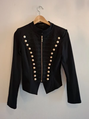 Svart rock från Merci Mademoiselle - Säljer en svart rock med coola dekorativa knappar i guld och broderade detaljer från Merci Mademoiselle. Jackan har lång ärm, rak passform och dragkedja framtill. Perfekt för dig som gillar statement-plagg och snygga detaljer.