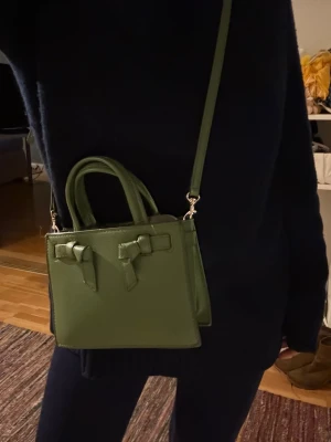 Grön axelväska från Zara - DISKUTERAR GÄRNA PRIS VID SNABB AFFÄR! Snygg grön axelväska från Zara med stilrena rosettdetaljer på framsidan. Väskan har både handtag och justerbar axelrem, samt dragkedja upptill. Tillverkad i syntetmaterial med en slät, lätt texturerad yta. Perfekt storlek för det viktigaste och ger en cool touch till din outfit.