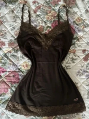 Brun linnetopp med spets från Hollister 2000s - Säljer en brun linnetopp från Hollister med tunna axelband och dekorativ spets i samma ton längs halsringning och nederkant. Toppen har en liten broderad Hollister-logga i hörnet och är gjord i mjukt material som känns skönt mot huden. Går inte att få tag på längre eftersom den kommer från 200-talet. ☺️💞// kan byta mot den zara knyt too jag söker 🤭