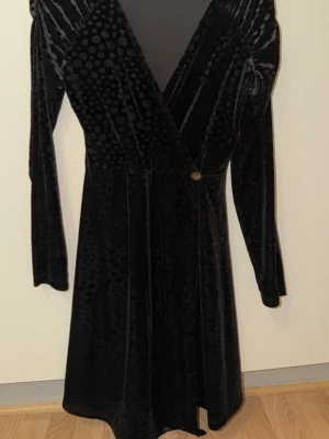 Svart omlottklänning i sammet - Snygg svart omlottklänning i mjuk sammet med prickigt mönster. Klänningen har lång ärm, markerad axel och en knapp i midjan. V-ringad och med skön passform, perfekt för dig som gillar lite extra detaljer.