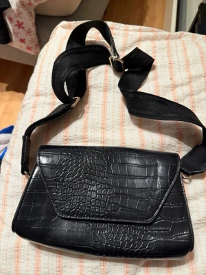 Svart axelväska med krokodilmönster - Snygg svart axelväska med krokodilmönstrad yta och bred justerbar axelrem i tyg. Väskan har ett magnetlås i guld och ett rymligt fack inuti. Perfekt för att bära med sig det viktigaste och ger en cool vibe till din outfit.