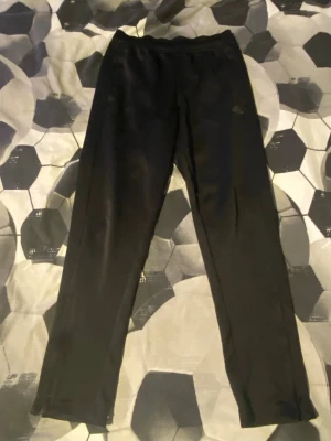 Svarta Adidas träningsbyxor M - Svarta träningsbyxor från Adidas med klassisk logga på vänster lår. Byxorna har elastisk midja och är tillverkade i mjukt syntetmaterial (Primegreen). Passar perfekt för sport och träning, med en slim passform och raka ben.