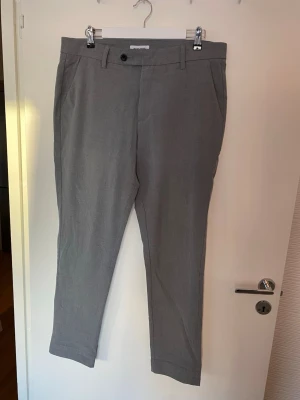 Grå kostymbyxor från Jack & Jones - Snygga grå kostymbyxor från Jack & Jones med klassisk passform och raka ben. Byxorna har bälteshällor, sidofickor och bakfickor med knapp. Materialet är mjukt och lätt, perfekt för en stilren look. Stängs med dragkedja och knapp framtill.