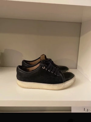 Lanvins - Säljer ett par svarta sneakers med snygg kombination av mocka och lackad tå. Skorna har rund tå, snörning och vit platt sula. Perfekta för dig som gillar stilrena och klassiska sneakers med en twist. Passar till många olika outfits.