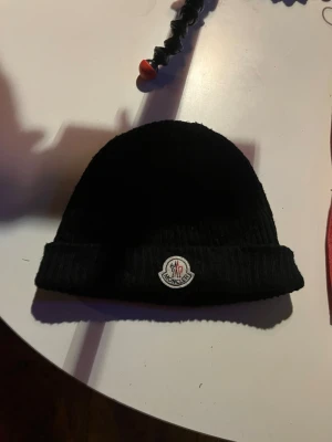 Svart Moncler mössa RIKTIG - Säljer en svart ribbstickad mössa från Moncler med klassisk logotyp framtill. Mössan är tillverkad i mjuk ull och har en uppvikt kant för extra stilpoäng. Perfekt för dig som vill ha en clean look och samtidigt hålla dig varm.