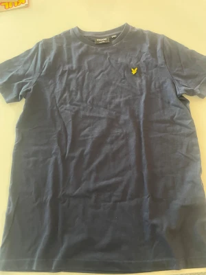 Marinblå t-shirt från Lyle & Scott - Snygg marinblå t-shirt från Lyle & Scott med klassisk rund hals och korta ärmar. T-shirten har en gul broderad logga på bröstet och är gjord i mjuk bomull som känns skön mot huden. Perfekt för dig som gillar stilrena och enkla plagg.