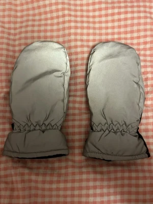 Reflex tumvantar - Säljer ett par reflex tumvantar där utsidan är silvrig/reflex medan insidan och handflatan är svart, och de har elastisk mudd vid handleden för extra komfort. Perfekta för kalla vinterdagar och håller händerna varma.