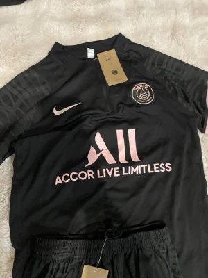 Svarta PSG träningskläder från Nike - Säljer ett svart PSG träningsset från Nike med rosa detaljer och logga på både tröja och byxor. Tröjan har halv dragkedja och lång ärm med mönstrade inslag. Byxorna har elastisk midja och rosa V-formad detalj. Perfekt för dig som älskar fotboll och vill ha snygga träningskläder.