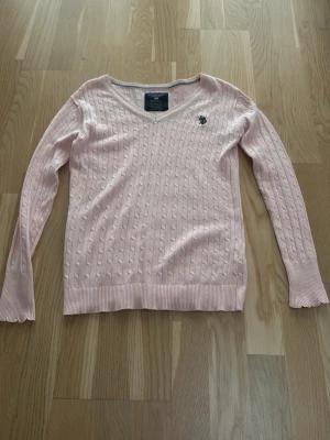 Ljusrosa kabelstickad tröja U.S. Polo Assn - Säljer en ljusrosa kabelstickad tröja från U.S. Polo Assn i storlek XS. Tröjan har v-ringad hals, långärmade ärmar med ribbade och vågiga muddar samt broderad logga på bröstet. Tillverkad i 100% bomull, perfekt för dig som gillar klassisk och stilren look. Det är inte ralhp louren men märket gick inte klika i det är u.s. Polo Assn
