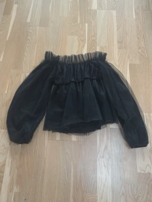 Svart offshoulder blus med volang - Säljer en svart blus med offshoulder-design och puffiga långärmar. Toppen har flera lager med volanger och är gjord i ett tunt, transparent syntetmaterial som ger en cool och luftig känsla. Perfekt för dig som gillar statement-plagg med extra volym.