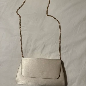 Champagnefärgad kuvertväska med kedja - Säljer denna kuvertväska i champagnefärgat material med en guldfärgad kedja som axelrem. I fint skick!💕