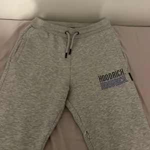 Hoodrich byxor XS  - Grå mjukisbyxor från Hoodrich i storlek XS. Byxorna har elastisk midja med snörning, sidofickor och en bakficka. Tryckt Hoodrich-logga i svart och blått på ena benet. Mjukt material som känns skönt mot huden, perfekt för chill dagar.