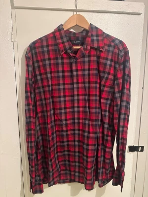 Rutig röd skjorta från Fred Perry - Säljer en snygg rutig skjorta från Fred Perry i storlek L. Skjortan har ett klassiskt röd-svart rutmönster, långa ärmar och knappar framtill. Perfekt för dig som gillar street och vill ha något som sticker ut lite extra.