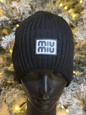 Svart ribbstickad mössa från Miu Miu - Svart ribbstickad mössa från Miu Miu med en tydlig logotyp framtill. Mössan är enkel men ändå snygg och passar perfekt för kalla dagar. Materialet känns mjukt och stretchigt, vilket gör den bekväm att bära.