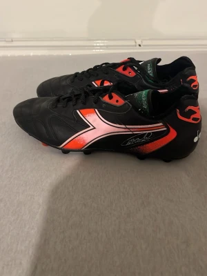 Diadora svarta och röda fotbollsskor - Säljer ett par Diadora fotbollsskor i svart med röda och rosa detaljer. Skorna har snörning, rund tå och platt sula med dobbar för gräsplan. Materialet är skinn och syntet, med en cool grafisk design på sidan och autograftryck. Perfekt för dig som vill sticka ut på planen. Van bastens skor och hans signatur på skorna