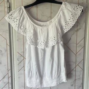 Vit blus med volang och broderade detaljer - Säljer en söt vit blus med bred volang runt axlarna och fina broderade blommor. Blusen är axelbandslös och har resår nertill. Superfint topp i storlek 170 men passar även xs och s