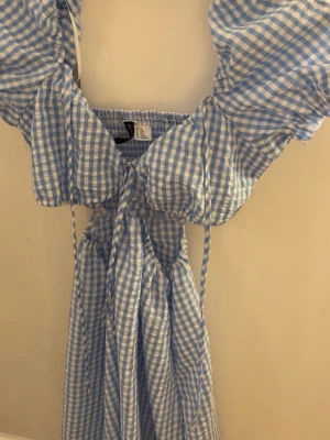 Blårutig klänning från H&M Divided - Supersöt blårutig kortklänning från H&M Divided med puffärmar, smock i ryggen och knyt fram. Tillverkad i tunn bomull, perfekt för varma dagar. Klänningen har en omlottkänsla och ger en riktigt somrig vibe.