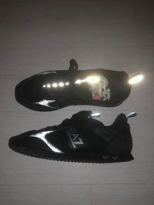 Svarta EA7 Emporio Armani sneakers - Säljer ett par svarta EA7 Emporio Armani sneakers med reflekterande detaljer och snörning. Skorna har en mix av mesh och syntetmaterial, samt logga på sidan. Snygg sportig look med platt sula och rund tå. Perfekta för dig som gillar stilrena och moderna sneakers.