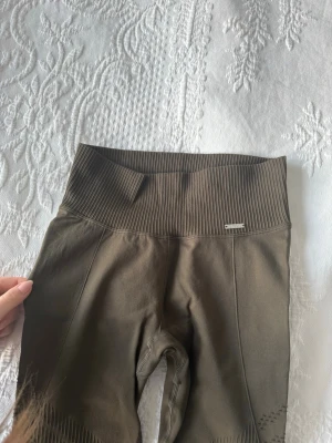 Mörkgrlna seamless leggings från Aim'n - Snygga bruna seamless leggings från Aim'n med ribbad hög midja och detaljer längs benen. Materialet är stretchigt och består av 92% polyamid och 8% elastan, vilket gör dem riktigt bekväma och perfekta för träning. Diskret Aim'n-logga framtill. Aldrig använda