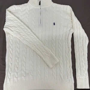 Vit stickad tröja Polo Ralph Lauren - Säljer en vit stickad tröja från Polo Ralph Lauren med klassiskt kabelmönster och halv dragkedja vid halsen. Tröjan har ribbade muddar och krage, samt den ikoniska blå Polo-loggan broderad på bröstet. Perfekt för kyliga dagar och snygg till jeans. Helt ny 