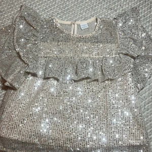 Glittrig blus med volang från Lindex - Säljer en glittrig blus från Lindex i storlek 146/152. Blusen är täckt av silverfärgade paljetter och har volangdetaljer över bröstet och på de korta ärmarna. Rund halsringning och knapp i nacken. Perfekt för dig som gillar att sticka ut!