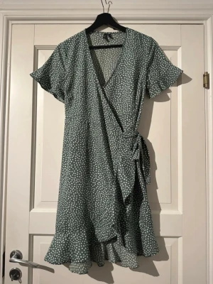 Grön prickig omlottklänning VeroModa - Superfin grön kort omlottklänning från VeroModa med vitt prickigt mönster. Klänningen har volangkant nertill, korta ärmar och knyt i midjan. V-ringad och lätt, perfekt för varma dagar. Materialet känns som mjuk polyester.