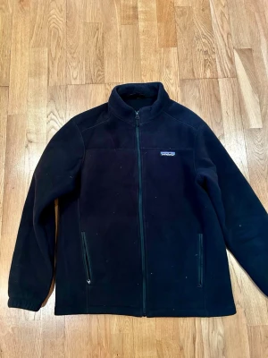 Svart fleecejacka från Patagonia - Svart fleecejacka från Patagonia i storlek M. Jackan har hel dragkedja framtill, två sidofickor med dragkedja och en hög krage. Perfekt för kyliga dagar och enkel att slänga på sig när det är lite kallare ute.