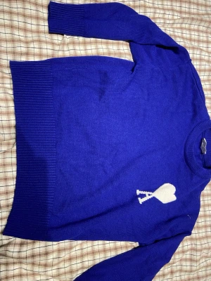 Blå stickad tröja från Ami Paris  - Snygg blå stickad tröja från Ami Alexandre Mattiussi med vitt hjärtmotiv på bröstet. Tröjan har rund hals, ribbade muddar och lång ärm. Perfekt för dig som gillar stilrena detaljer och vill ha något som sticker ut lite extra.