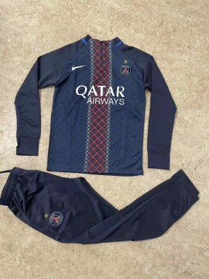 PSG träningsset från Nike - Säljer ett mörkblått PSG träningsset från Nike med långärmad tröja och matchande byxor. Tröjan har rött och vitt rutmönster framtill, Qatar Airways-logga och PSG-emblem. Byxorna har PSG-logga och resår i midjan. Perfekt för fotbollsträning.