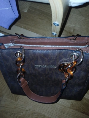 Brun handväska Michael Kors - Snygg brun handväska från Michael Kors med MK-logga över hela väskan. Väskan har guldfärgade och bruna ringdetaljer på handtaget, dragkedja upptill och ett stort fack med ljus insida. Perfekt storlek för allt du behöver och stilren design.