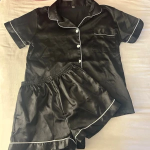 Svart sidenpyjamas med vit kant - Säljer en svart pyjamas i siden med vit piping längs kanterna. Setet består av en kortärmad skjorta med knappar och bröstficka samt matchande shorts med resår i midjan. Inte använd mycket.