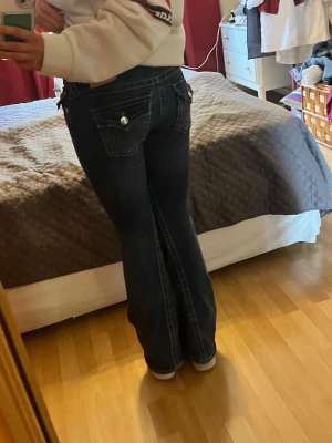 True Religion Joey Low Rise Flare Jeans - Säljer ett par mörkblå True Religion Joey jeans med låg midja och flare-ben. Snygga detaljer med kontrastsömmar och stora fickor med knappar bak. Klassisk jeanslook med coola broderier och slitstarka material. Perfekt för dig som gillar statement-jeans! 