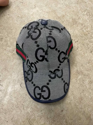 Grå och svart GG-mönstrad Gucci keps - Snygg keps från Gucci med GG-logga i svart och grått över hela kepsen. Har klassiska gröna och röda ränder på sidorna och justerbar rem baktill. Materialet är troligtvis bomull med detaljer i syntet. Perfekt för dig som vill sticka ut med en ikonisk accessoar.