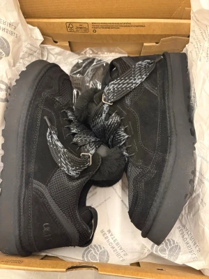 Svarta sneakers från UGG - Säljer ett par svarta UGG sneakers med fluffig päls runt kanten och grova snören i svart och grått mönster. Skorna har en robust platt sula, rund tå och detaljer i mocka och mesh. Perfekta för dig som vill ha både stil och komfort under kyliga dagar. Aldrig använda pga va för stora. Extra snöre följer även med. Priset kan diskuteras!