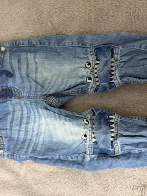 Barnjeans med hajdetaljer – stl 90 - Söta och coola jeansbyxor för barn i storlek 90 med roligt hajmotiv på knäna. Mjuka och bekväma med resår i midjan och snörning, perfekt för lek och vardag. Använda men i fint skick, inga hål eller fläckar.