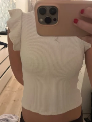 Vit ribbad topp med volangärm - Supersöt vit ribbad topp med volangärmar och rund halsringning. Toppen är croppad och har en tight passform som framhäver figuren. Perfekt att styla med jeans eller kjol för en fräsch look.