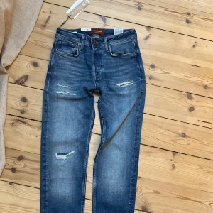 Blå jeans från Jack & Jones, storlek 28/32 - Snygga blå jeans från Jack & Jones med slitna detaljer och klassisk femficksdesign. Jeansen har slim passform,  och är tillverkade i jeansmaterial med lite stretch. Perfekt för dig som gillar en avslappnad och trendig look.