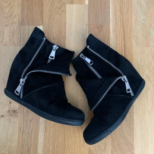 Svarta boots med dragkedjor och kilklack - Unika svarta boots med kilklack och stora silvriga dragkedjor som detaljer. Skorna har en rund tå och är tillverkade i mocka för en mjuk och cool känsla. Perfekt för dig som gillar edgy stil och vill sticka ut med dina skor.