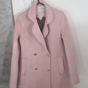 Rosa kappa från Rinascimento XS - En vacker rosa kavaj/kappa från Rinascimento i storlek XS. Denna kappa har en dubbelknäppning med guldknappar och en klassisk krage. Denna kavaj är verkligen fin och skicket är fint, men tyvärr finns det upptill på baksidan en inte så tydlig fläck. På bilden framkommer fläcken tydligare, men i verkligheten syns inte fläcken tydligt. Måttet rakt över midjan är ca 42 cm, från armhåla till armhåla är måttet ca 45 cm och längden är ca 83 cm.