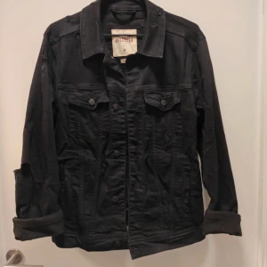 Svart jeansjacka från Hollister M - Svart jeansjacka från Hollister i stretchigt material. Klassisk modell med krage, två bröstfickor med knappar och knappstängning framtill. Jackan har långa ärmar och uppvikta muddar för en chill vibe. Perfekt för dig som gillar streetstyle.