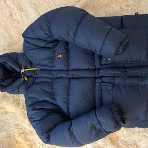 Mörkblå jacka från Fjällräven - Säljer en mörkblå dunjacka från Fjällräven med huva och snygga gula detaljer. Jackan är vadderad med 90% gåsdun och 10% fjäder, yttertyg och foder i polyamid. Klassisk pufferstil med tryckknappar och dragkedja framtill.