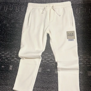 Vita mjukisbyxor från Burberry - Snygga vita mjukisbyxor från Burberry med ribbad struktur och dragsko i midjan. Byxorna har raka ben, två fickor fram och en diskret Burberry-logga på ena benet. Perfekta för en chill och stilren look.
