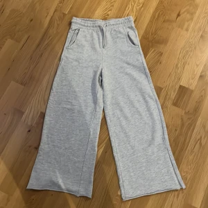 Grå wide mjukisbyxor från KappAhl - Snygga ljusgrå mjukisbyxor från KappAhl i storlek 146. Byxorna har breda ben, slits vid benslut, elastisk midja med snörning och två fickor fram. Perfekta för chill dagar och riktigt sköna tack vare mjukt bomullsmaterial.