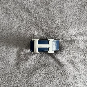 Hermès bälte  - Säljer ett stilrent Hermès bälte med ikoniskt H-spänne i silverfärgad metall. Ena sidan är marinblå med vit söm, andra sidan är svart. Bältet är tillverkat i äkta skinn och har en klassisk, rak form. Perfekt för dig som vill ha en lyxig accessoar med tydlig designer-vibe.