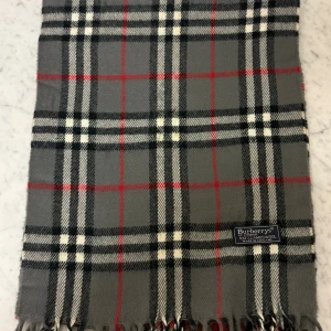 Rutig grå halsduk från Burberrys - Snygg halsduk från Burberrys i klassiskt rutigt mönster med grå, svart, vit och röda detaljer. Gjord i 100% lammull, mjuk och varm med fransar i kanten. Perfekt accessoar för att lyfta din stil under kyliga dagar.