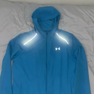 Blå vindjacka från Under Armour - Blå vindjacka från Under Armour med huva, hel dragkedja och reflekterande detaljer på axlarna. Jackan har logga på bröstet och stor text på ryggen. Tillverkad i lätt polyester, perfekt för träning utomhus. Snygg och sportig look.