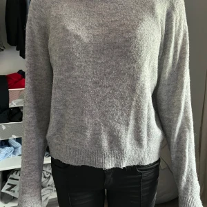 Grå stickad tröja med rund hals - Mysig grå stickad tröja med rund hals och ribbade muddar vid ärmslut och nederkant. Modellen är lite loose och har långa ärmar, perfekt för att hålla sig varm under kyliga dagar. Enkel och stilren design som funkar till många olika outfits.