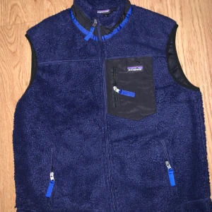 Blå fleeceväst från Patagonia - Säljer en mörkblå fleeceväst från Patagonia med svarta detaljer och blå dragkedjor. Västen har tre fickor med dragkedja, varav en bröstficka med logga. Perfekt för lager-på-lager och outdoor. Snygg, sportig look med hög krage.