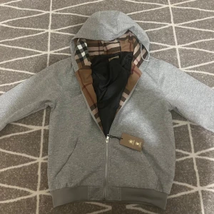 Grå zip up hoodiejacka från Burberry Med dragkedja  - Säljer en grå hoodiejacka från Burberry med klassiskt rutigt foder i huvan. Jackan har dragkedja framtill, ribbade muddar och två fickor. Perfekt för dig som gillar stilrena och exklusiva plagg med en twist av ikoniskt mönster.
